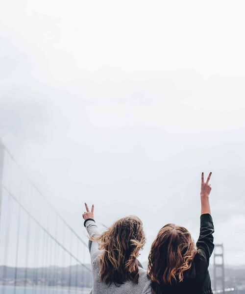 Zwei Personen stehen Rücken an Rücken mit erhobenen Armen und zeigen Peace-Zeichen vor einer nebeligen Brücke.