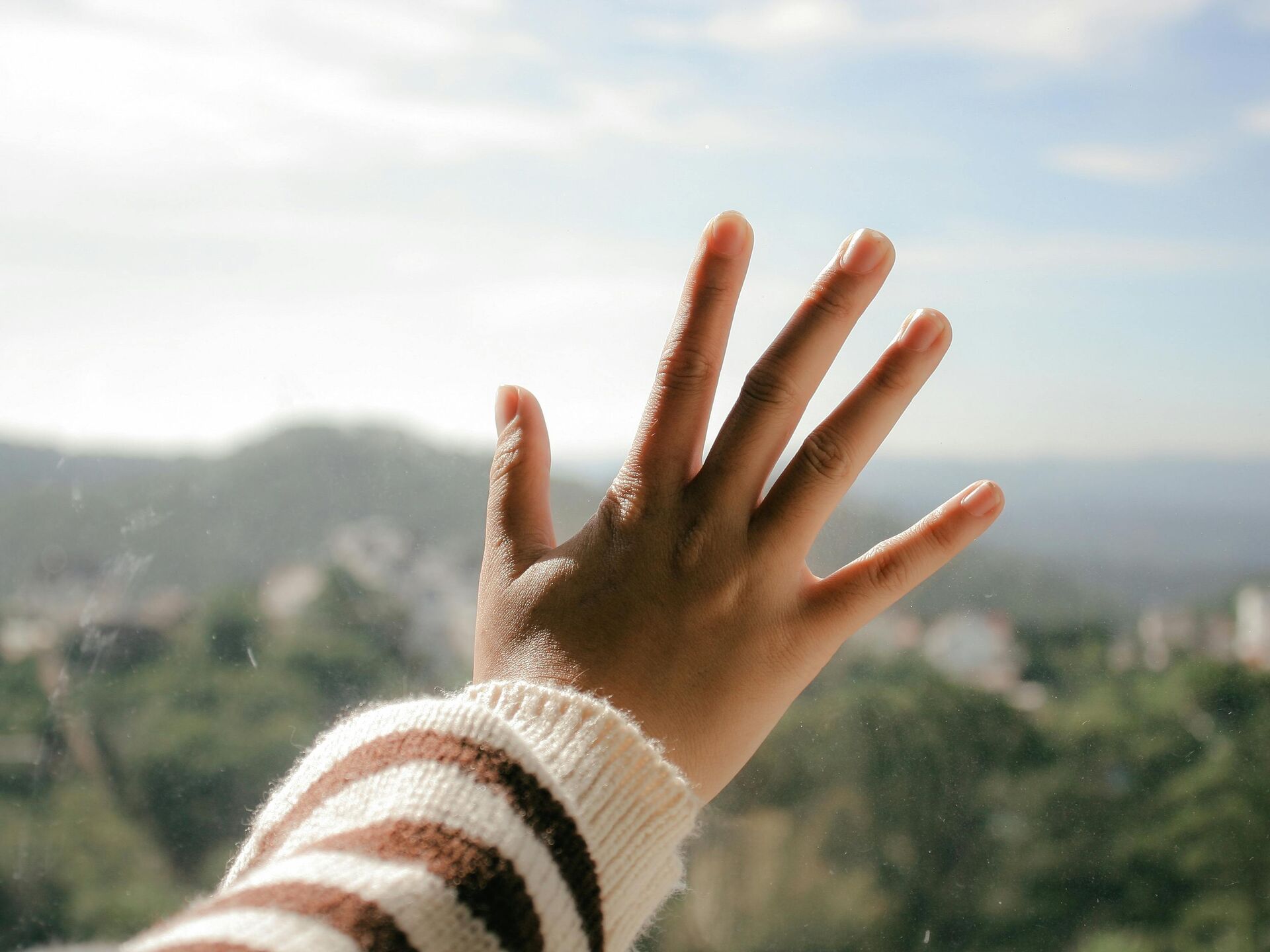 Hand mit gestreiftem Pulloverärmel, die sich an ein Fenster legt, im Hintergrund eine verschwommene Landschaft.