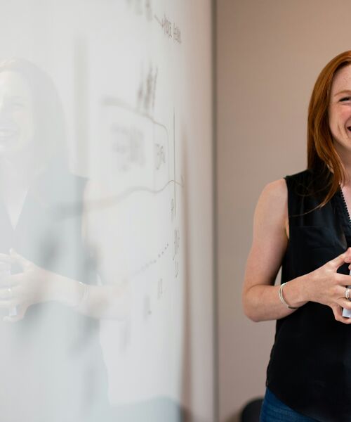 Lächelnde Frau steht vor einem Whiteboard und erklärt ein Thema in einem modernen Seminarraum.