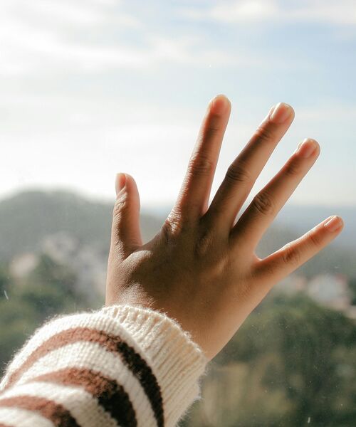 Hand mit gestreiftem Pulloverärmel, die sich an ein Fenster legt, im Hintergrund eine verschwommene Landschaft.