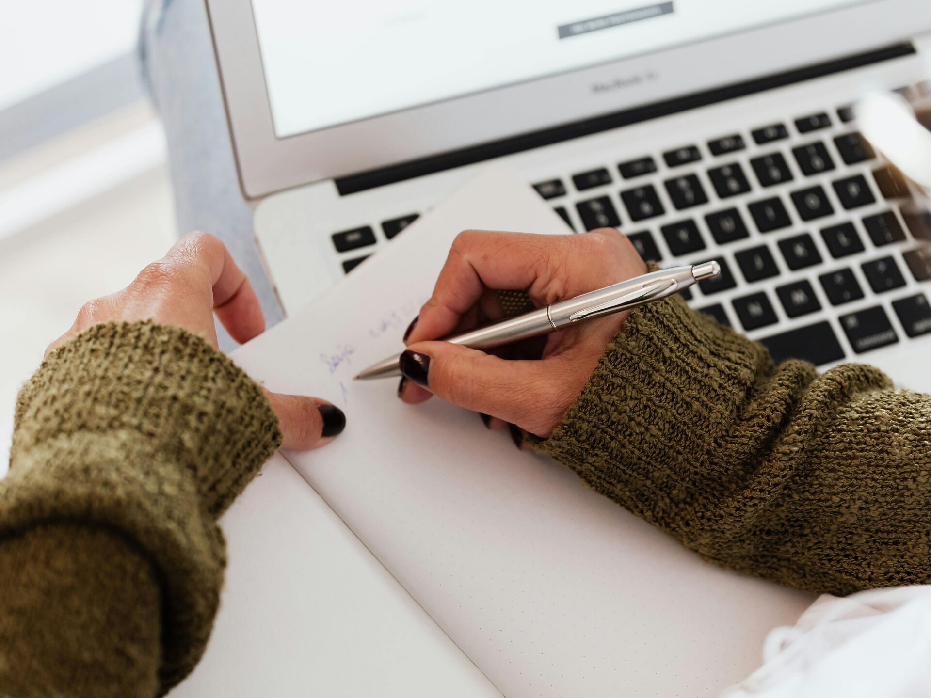 Hand mit grünem Strickpullover schreibt mit einem silbernen Stift in ein Notizbuch vor einem geöffneten Laptop.