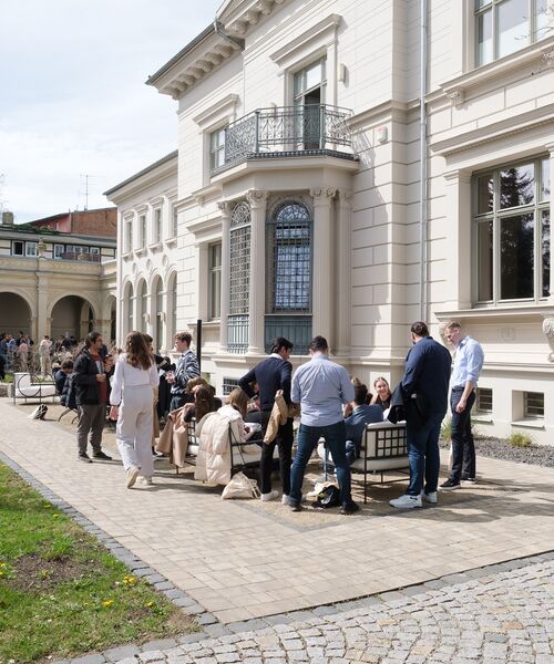 Gruppe von Menschen sitzt und steht vor einem historischen Gebäude im Freien – Symbol für Austausch und gemeinsames Lernen in angenehmer Atmosphäre.