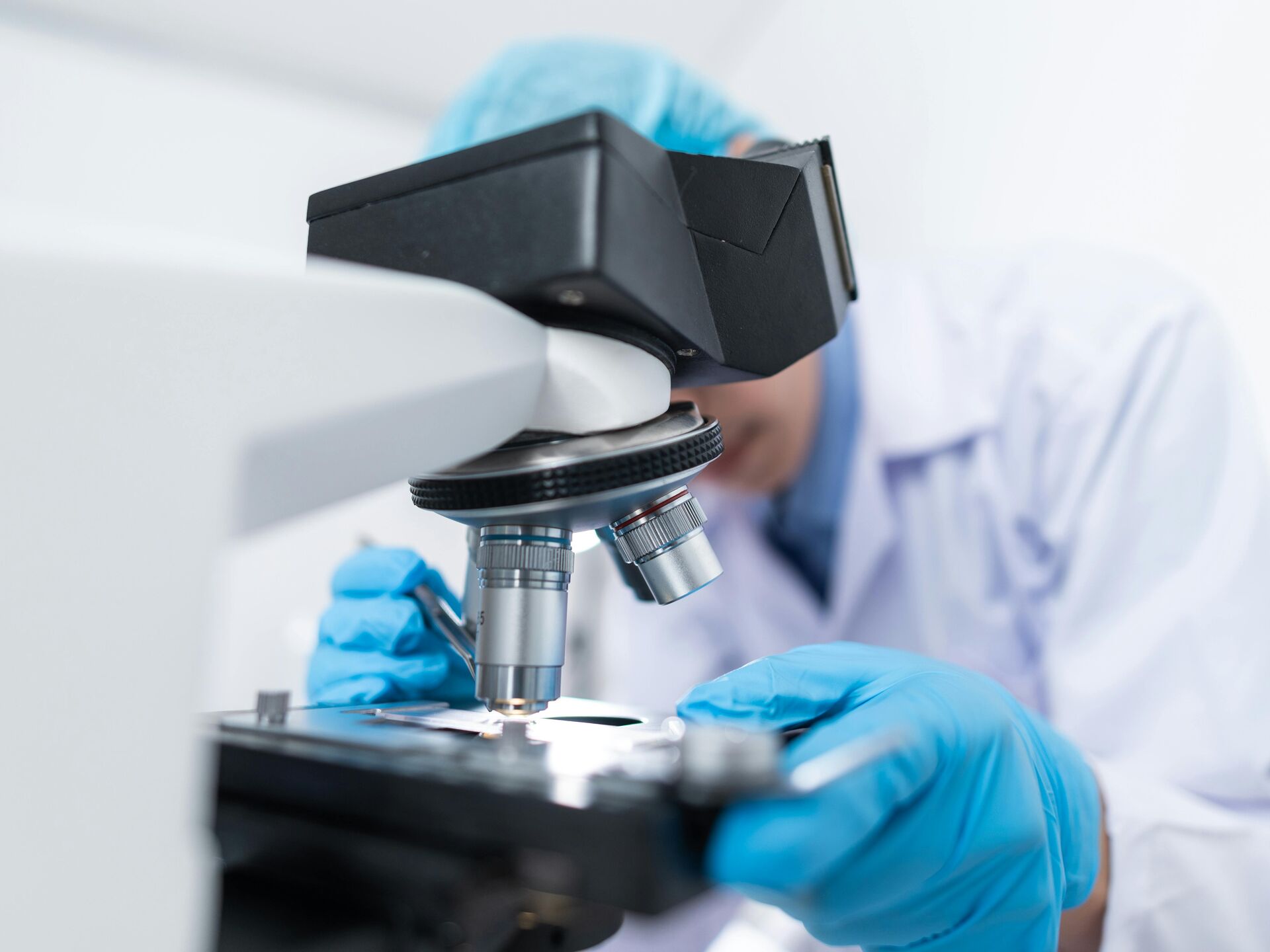 Person mit blauen Handschuhen arbeitet im Labor und betrachtet eine Probe unter dem Mikroskop – Symbol für Forschung und wissenschaftliche Analyse.