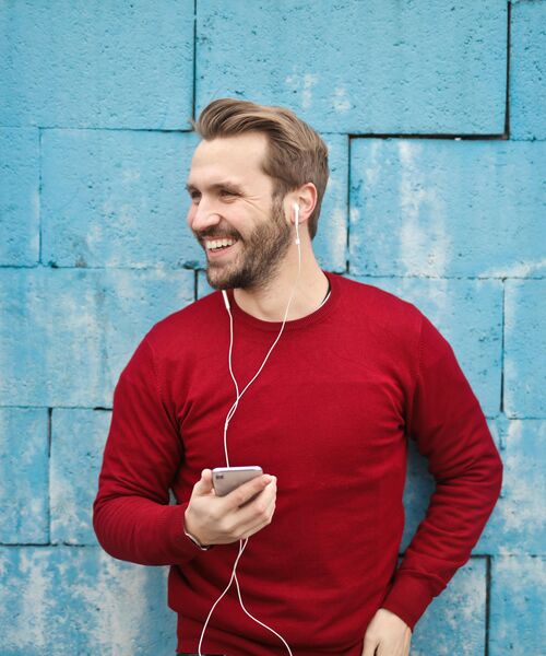 Lächelnde Person lehnt an einer blauen Wand, hält ein Smartphone in der Hand und hört Musik über weiße Kopfhörer.