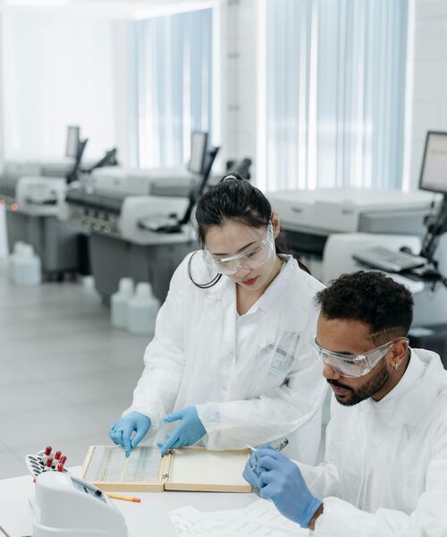 Zwei Personen in Laborkleidung und Schutzbrillen arbeiten gemeinsam an Unterlagen – Symbol für wissenschaftliche Zusammenarbeit und Forschung.