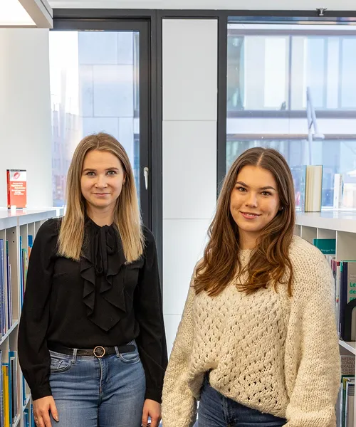 Zwei Frauen sitzen in einer hellen Bibliothek zwischen Bücherregalen und lächeln in die Kamera.