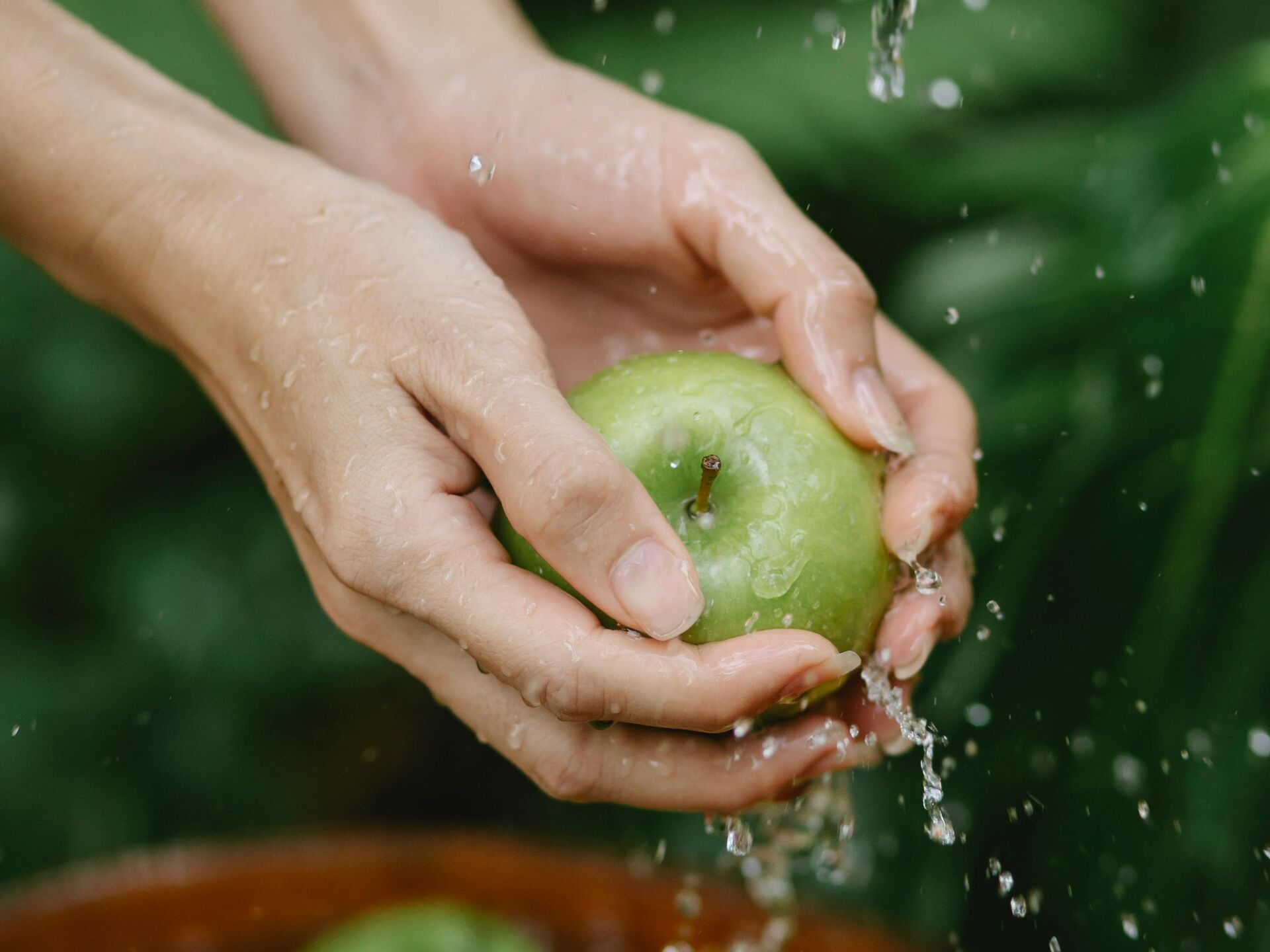 Hände waschen einen grünen Apfel unter fließendem Wasser, weitere Äpfel liegen in einer Schale darunter.