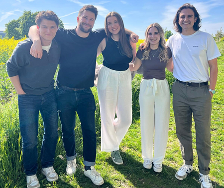 Fünf junge Menschen stehen lachend im Grünen und genießen bei Sonnenschein den Tag.