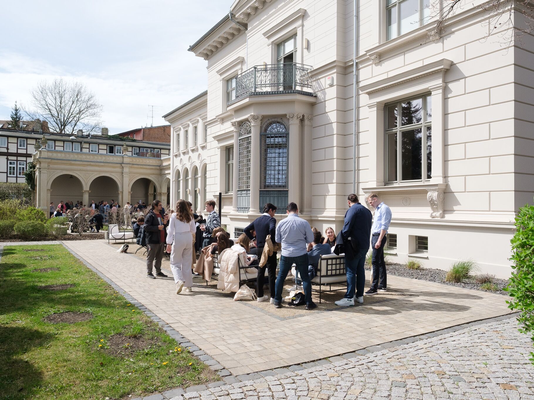 Gruppe von Menschen sitzt und steht vor einem historischen Gebäude im Freien – Symbol für Austausch und gemeinsames Lernen in angenehmer Atmosphäre.