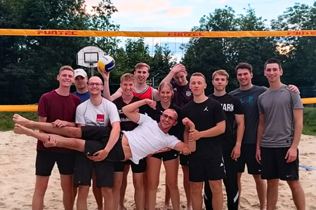 Gruppe junger Menschen steht lachend auf einem Beachvolleyballfeld, eine Person wird spaßig hochgehoben.