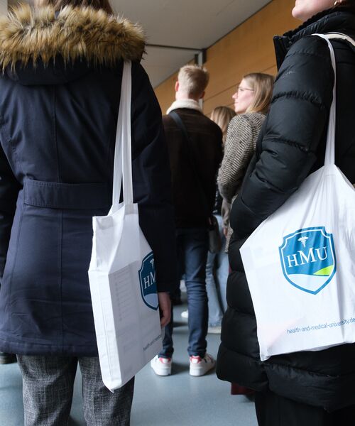 Mehrere Personen stehen zusammen, zwei tragen weiße Stofftaschen mit dem Logo der HMU – Health and Medical University.