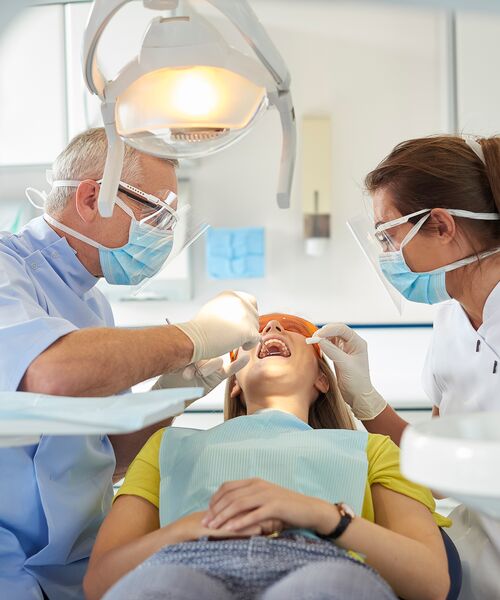 Zahnarzt und Assistentin behandeln eine Patientin auf dem Behandlungsstuhl unter einer hellen Lampe – Symbol für professionelle zahnärztliche Versorgung.