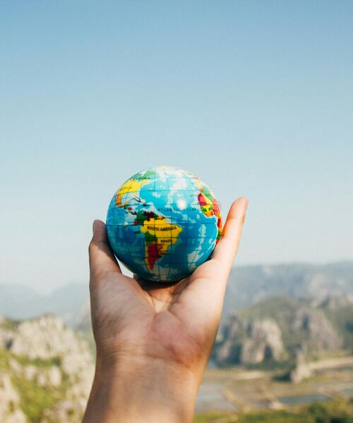 Hand hält einen kleinen Globus vor einer Landschaft mit Bergen und blauem Himmel.