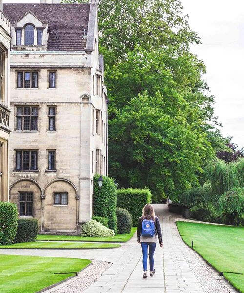 Person mit Rucksack geht einen Weg entlang zwischen historischem Gebäude und gepflegtem Garten.