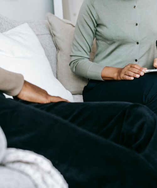 Zwei Personen sitzen auf einem Sofa, eine hält ein Tablet in der Hand, die andere sitzt entspannt gegenüber.
