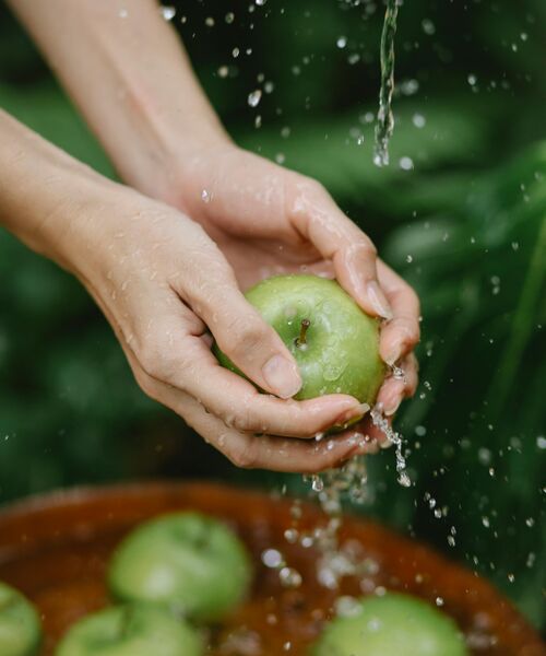 Hände waschen einen grünen Apfel unter fließendem Wasser, weitere Äpfel liegen in einer Schale darunter.