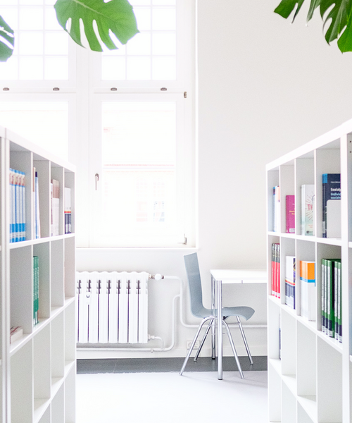 Heller Bibliotheksraum mit Bücherregalen, einem Tisch und einem Stuhl vor einem großen Fenster.