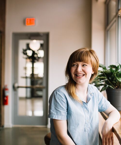 Junge Frau sitzt lächelnd an einem Fensterplatz in heller Umgebung, wirkt entspannt und freundlich.