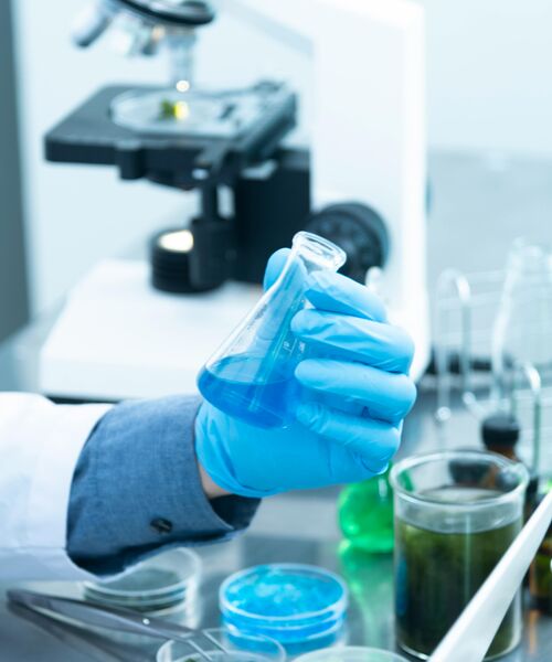 Hand mit blauem Schutzhandschuh hält ein Becherglas mit blauer Flüssigkeit, im Hintergrund Laborgeräte – Symbol für wissenschaftliche Forschung und Experimente.