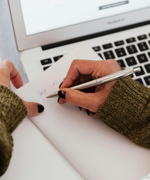Hand mit grünem Strickpullover schreibt mit einem silbernen Stift in ein Notizbuch vor einem geöffneten Laptop.