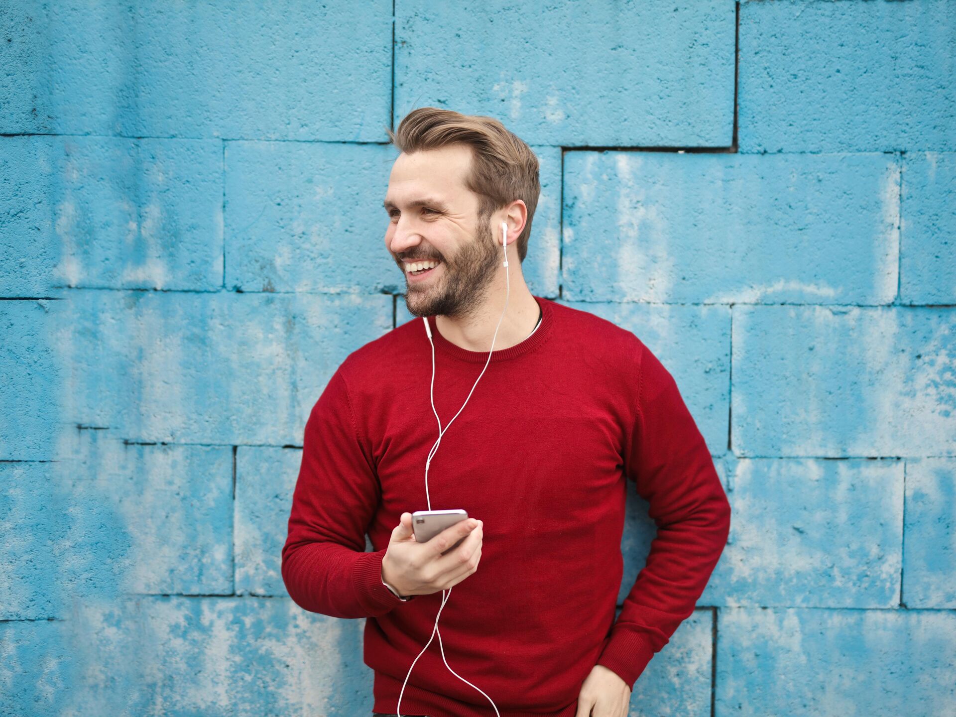 Lächelnde Person lehnt an einer blauen Wand, hält ein Smartphone in der Hand und hört Musik über weiße Kopfhörer.
