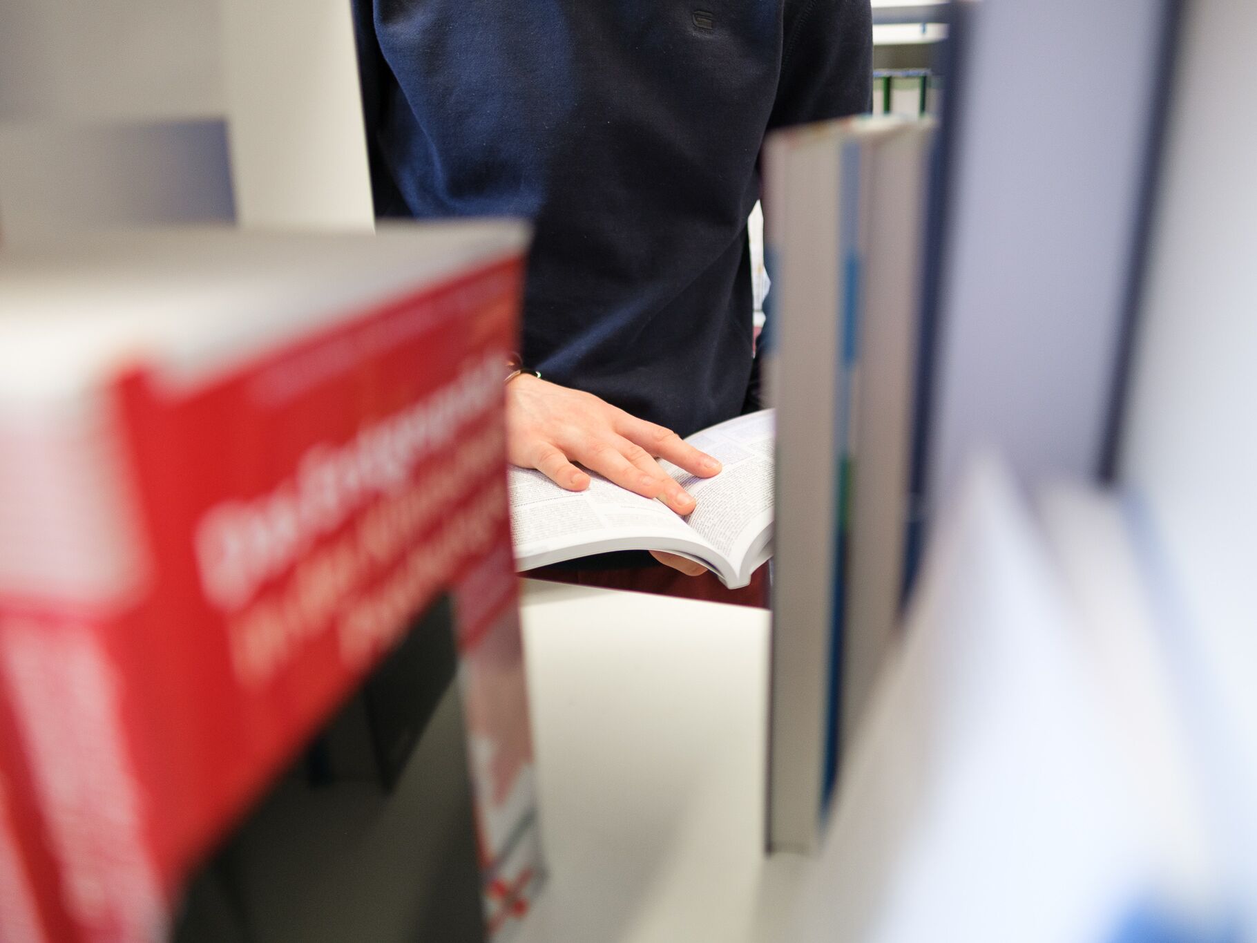Person blättert in einem aufgeschlagenen Buch zwischen Bücherregalen in einer Bibliothek.