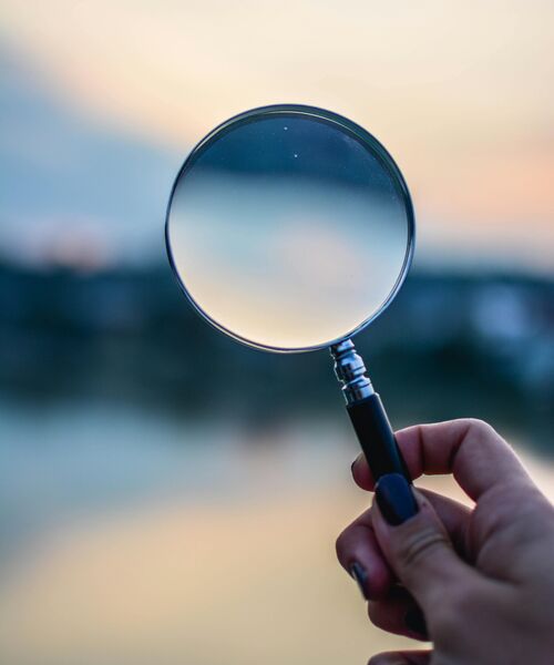 Hand hält eine Lupe vor unscharfem Hintergrund – Symbol für Suche, Analyse oder genaues Hinsehen.