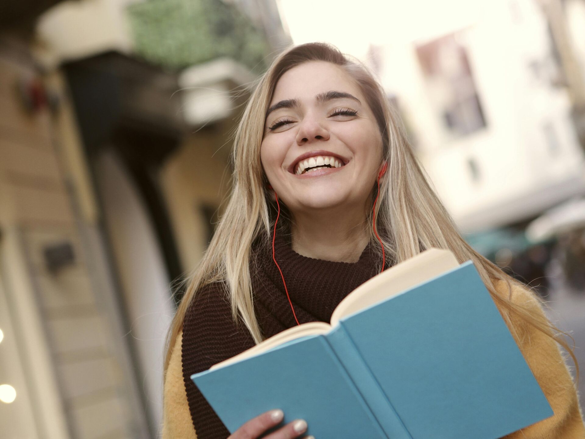 Lachende junge Frau mit Kopfhörern hält ein geöffnetes Buch in der Hand – Symbol für Freude am Lernen und Wissen.