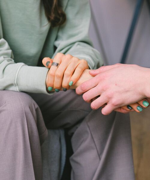 Zwei Personen sitzen einander gegenüber, eine legt beruhigend die Hand auf die der anderen – Symbol für Unterstützung und Vertrauen.